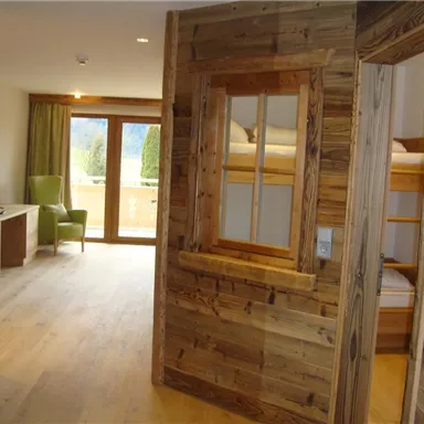 A modern furnished room with wooden paneling and large windows. In the background, a cozy armchair and a bunk bed can be seen.