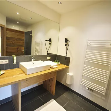 A modern bathroom with a wooden vanity and a large mirror. The walls are light and there are elegant dark tiles.
