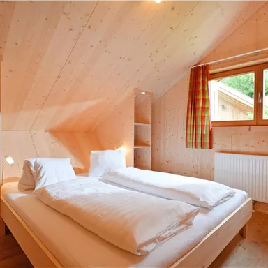 A cozy bedroom with a double bed and light wood paneling. Large windows let in plenty of daylight.