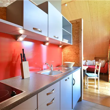 A modern kitchen with a red wall, bright cabinets, and a sink. In the background, a table and chairs are visible.