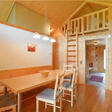 A cozy room with a wooden ceiling and a dining table. A staircase leads to an upper area.