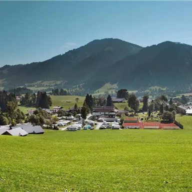 An idyllic landscape with green meadows and mountains in the background. In the foreground, there are some buildings and tennis courts visible.