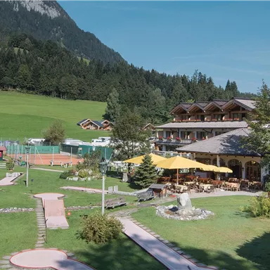 A picturesque landscape with a hotel and well-kept gardens. In the background, green hills and trees can be seen.