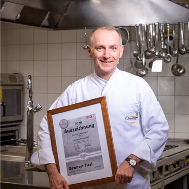 A chef in a professional kitchen proudly holds an award in his hand. In the background, kitchen equipment and pots are visible.