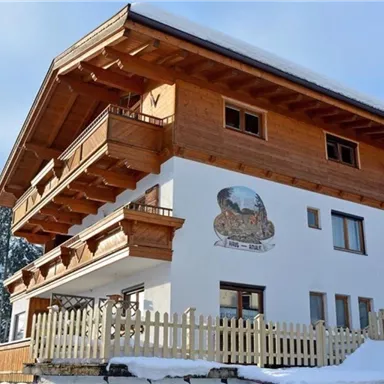 A cozy chalet in alpine style with wooden balconies. Surrounded by snow-covered trees and a clear winter atmosphere.
