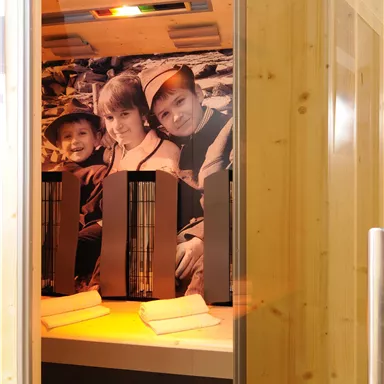 An interior of a sauna with wooden paneling and a picture of three smiling children on the wall. In the foreground, two heat lamps are on a bench.