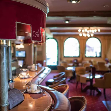 Eine gemütliche Restaurant-Innenansicht mit einer Holztheke und hellen Fenstern. Rote Teppiche und bequeme Stühle schaffen eine einladende Atmosphäre.
