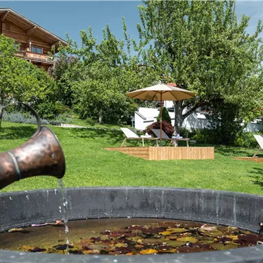Ein hübscher Garten mit einem Teich und einem Wasserkrug im Vordergrund. Im Hintergrund stehen Liegen und Sonnenschirme unter einem Baum.