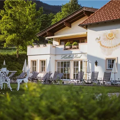 Ein charmantes Haus mit einem hellen Außenbereich und eleganten Liegen. Die grüne Umgebung und die Berge im Hintergrund schaffen eine entspannende Atmosphäre.