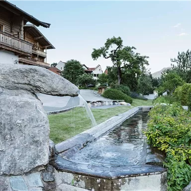 Ein ruhiger Garten mit einem steinernen Wasserlauf. Im Hintergrund sind Holzhäuser und grüne Bäume zu sehen.