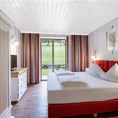 Ein modernes Schlafzimmer mit einem roten Bett und zwei Kissen. Große Fenster bieten einen Blick auf die grüne Landschaft.