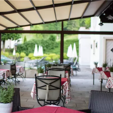 Ein gemütlicher Restaurantbereich mit gedeckten Tischen und Stühlen. Im Hintergrund sind grüne Landschaften und Sonnenschirme zu sehen.