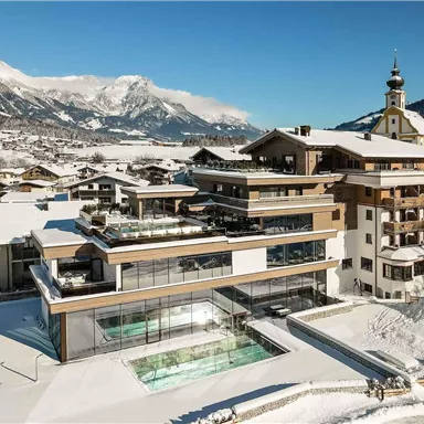 A modern hotel building in a snowy mountain landscape. In the foreground, there is a pool surrounded by snow-covered areas.