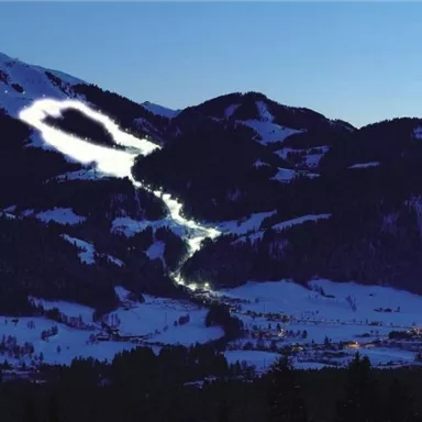 A snow-covered mountain landscape at night with illuminated slopes. In the background, dark mountains and a clear sky can be seen.