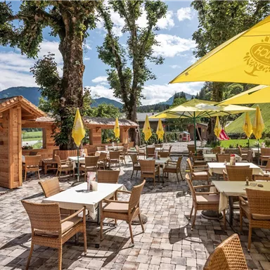A cozy outdoor restaurant with tables and chairs. Yellow umbrellas provide shade on a sunny day.