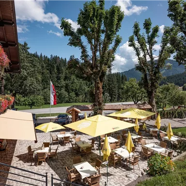An inviting restaurant with numerous outdoor tables and yellow umbrellas. In the background, green trees and a clear sky can be seen.