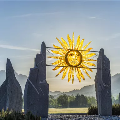 Eine Gruppe von großen Steinen bildet einen Rahmen für eine strahlende Sonne im Hintergrund. Die Landschaft zeigt sanfte Hügel und einen klaren Himmel.