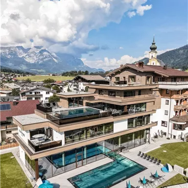 A modern hotel with a clear outdoor pool and loungers. In the background, mountains and a blue sky can be seen.