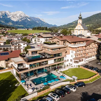 A modern hotel with a pool and beautiful garden facility. In the background, the mountains and a church can be seen.