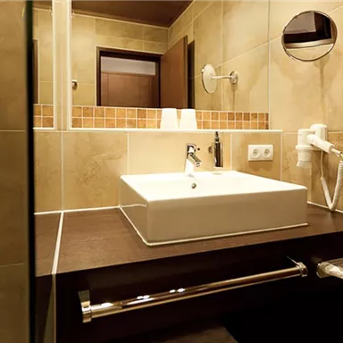 A modern bathroom with a bright sink, wooden shelf, and beige tiled walls. The room is well-lit and clean.