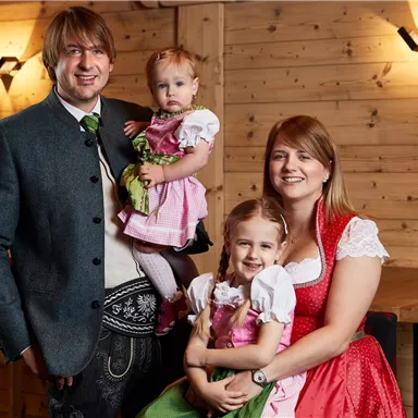 A family in traditional costumes stands in a cozy wooden room. The adults and children smile warmly at the camera.