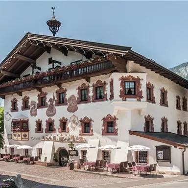 A traditional Bavarian building with a beautiful view of the mountains. In front of the house, tables with sun umbrellas are set up.