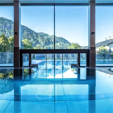 A modern indoor swimming pool with large windows and views of the mountains. The water surface reflects the surroundings and creates a relaxing atmosphere.