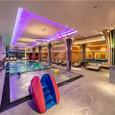A modern indoor pool with colorful lights and comfortable loungers. In the foreground, there is a play area for children with a slide.