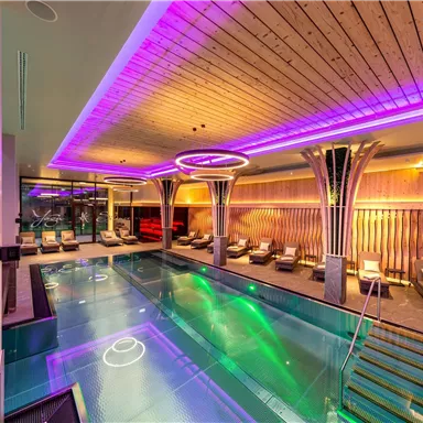 An elegant indoor pool with colorful LED lights and modern loungers. The relaxing atmosphere is enhanced by the stylish wooden ceiling and soft colors.