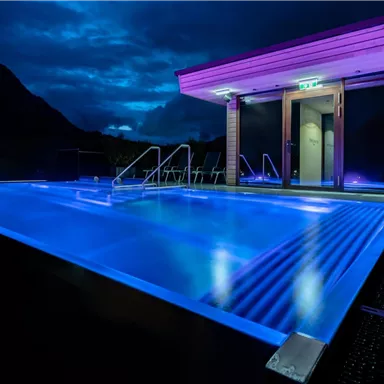 A modern pool with blue lights in a tranquil setting. The sky is overcast and there is stylish architecture in the background.