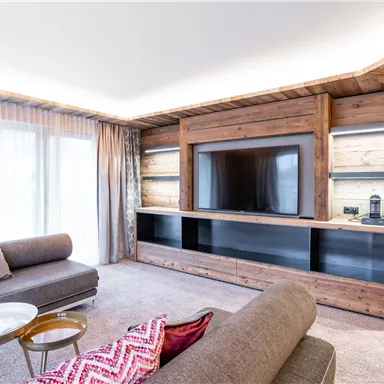 A modern living room with wooden walls and a large TV. There are comfortable sofas and decorative pillows in pink.
