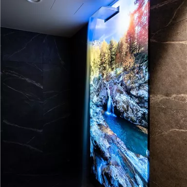 A modern bathroom with a large digital screen displaying a waterfall scene. The walls are made of dark stone, creating an elegant contrast to the colorful landscape.