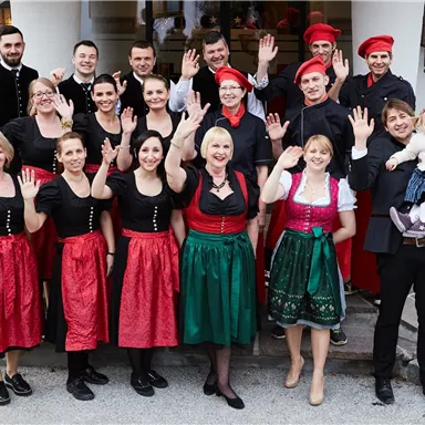 A group of people in traditional clothing smiles and waves. They stand in front of a building and appear friendly and inviting.