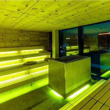A modern sauna interior with wooden shelves and soft green lighting. Large windows offer a view outside and create a relaxing atmosphere.