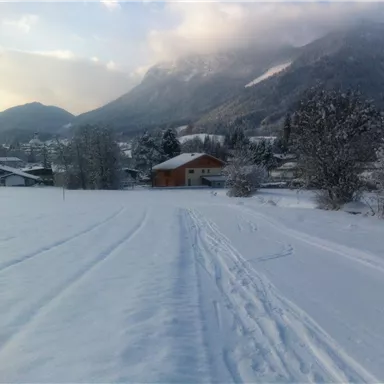 A snowy landscape with hills in the background. A cozy house stands nearby, surrounded by trees.