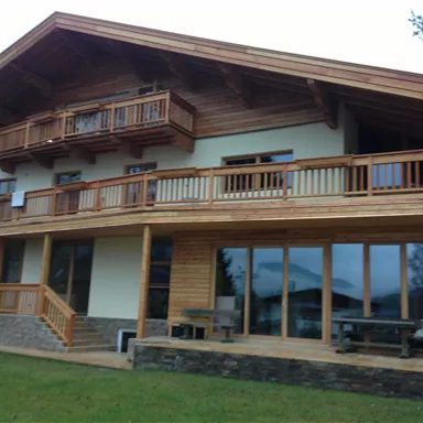 A modern wooden house with a spacious balcony and large windows. The terrace is surrounded by green grass.