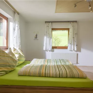 Ein gemütliches Schlafzimmer mit einem großen Bett und bunten Kissen. Große Fenster sorgen für viel Tageslicht und einen Blick ins Grüne.