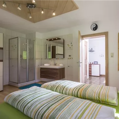 Ein modernes Schlafzimmer mit zwei Einzelbetten, die mit gestreiften Bettdecken bedeckt sind. Im Hintergrund befinden sich ein Badezimmer und ein heller Eingangsbereich.