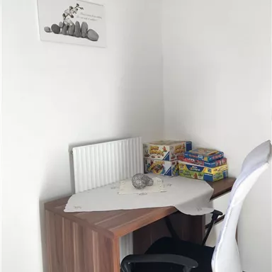 Ein einfaches Arbeitszimmer mit einem Holztisch und einem weißen Stuhl. Auf dem Tisch befindet sich eine Tischdecke, ein Stein und einige Spiele.