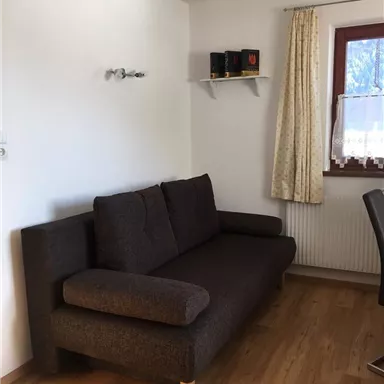 Ein gemütliches Wohnzimmer mit einem braunen Sofa und einem Fenster. An der Wand befindet sich ein Regal mit Dekorationen.