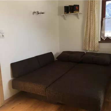 Ein schlichtes Zimmer mit einem umgebauten Sofa und einem Fenster, das Tageslicht hereinlässt. Die Wände sind hell und es gibt eine kleine Regalfläche.