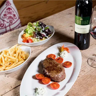 A delicious dish with steak, fresh tomatoes, fries, and a colorful salad. Accompanied by a glass of red wine on a wooden table.