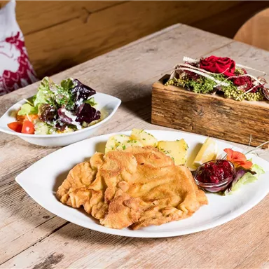 A plate with breaded meat, potatoes, and red sauce, next to a fresh salad with various vegetables. The table is made of light wood, and in the background, there is a decorative flower box.