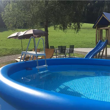 Ein aufblasbarer Pool mit einer Leiter steht in einem grünen Garten. Neben dem Pool sind eine Rutsche, Liegen und Sonnenschirme zu sehen.