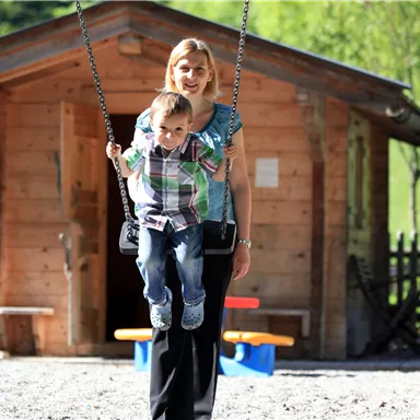 A woman swings a child on a swing in a playground. In the background, a wooden hut can be seen.