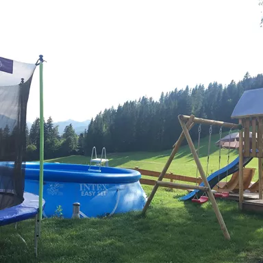 Ein Spielbereich mit einem Trampolin, einem Swimmingpool und einem Spielplatz. Im Hintergrund sind grüne Wiesen und Bäume zu sehen.