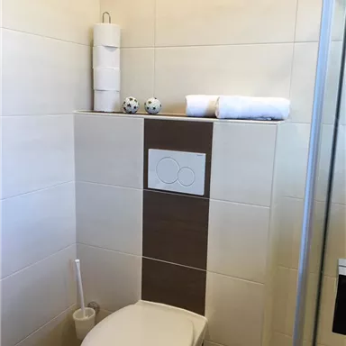 Ein modernes Badezimmer mit einer weißen Toilette und einem braunen Wandakzent. Saubere Handtücher und dekorative Gegenstände sind auf einer Ablage platziert.