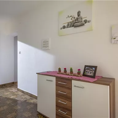 Ein heller Flur mit einem weißen Schrank und einer Holzoberfläche. Auf einer pinken Tischdecke stehen kleine Pflanzen und Bilderrahmen.