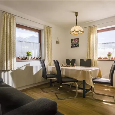 Ein gemütlicher Essbereich mit einem Tisch und Stühlen. Große Fenster sorgen für viel Tageslicht und Pflanzen dekorieren die Fensterbank.