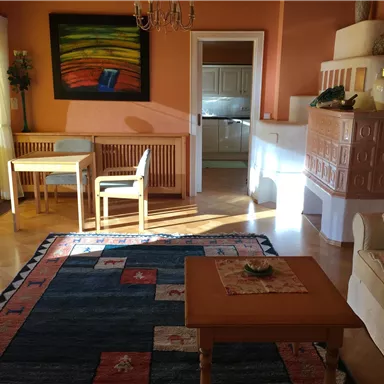 A cozy living room with a carpet, a dining table, and chairs. The walls are painted in warm colors, and there is an open kitchen in the background.